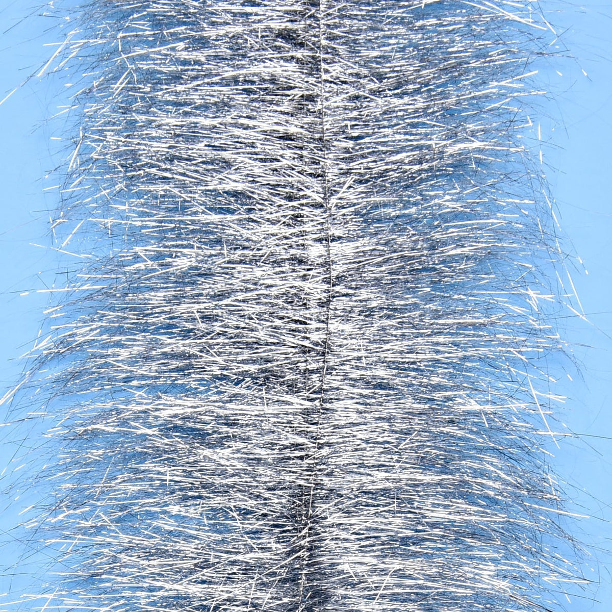 Silver metallic tinsel on dense sparkle brush for fly tying, shimmering strands against pale blue background