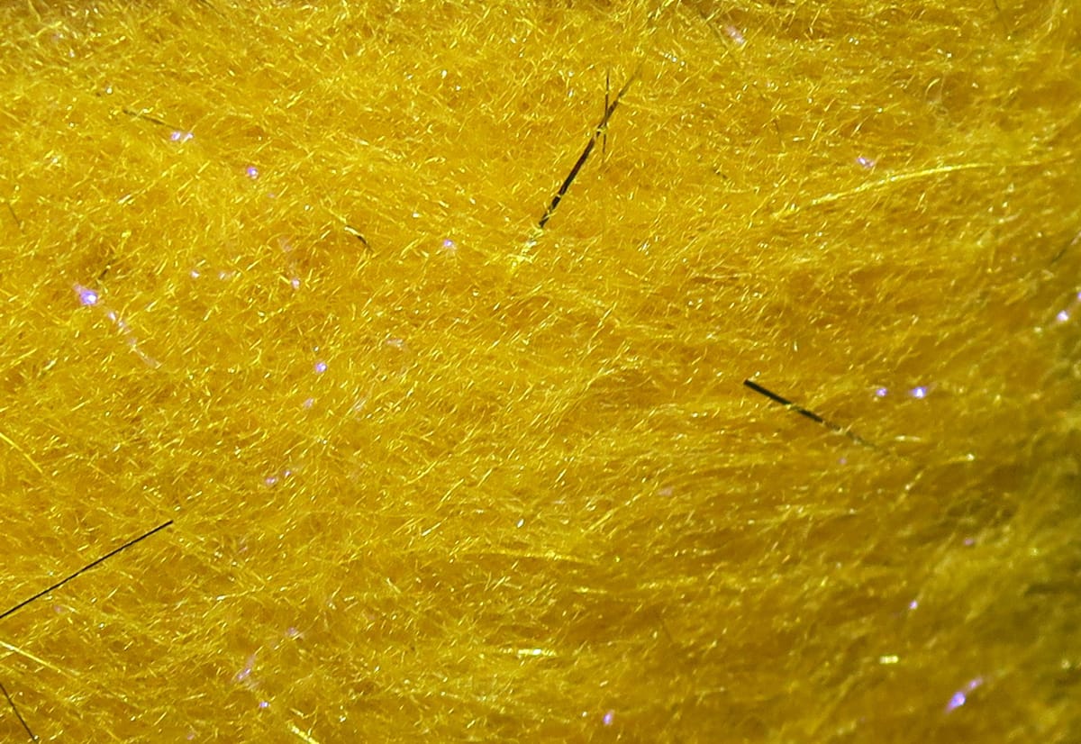Bright golden yellow synthetic dubbing fibers with iridescent flash and scattered black guard hairs for fly tying
