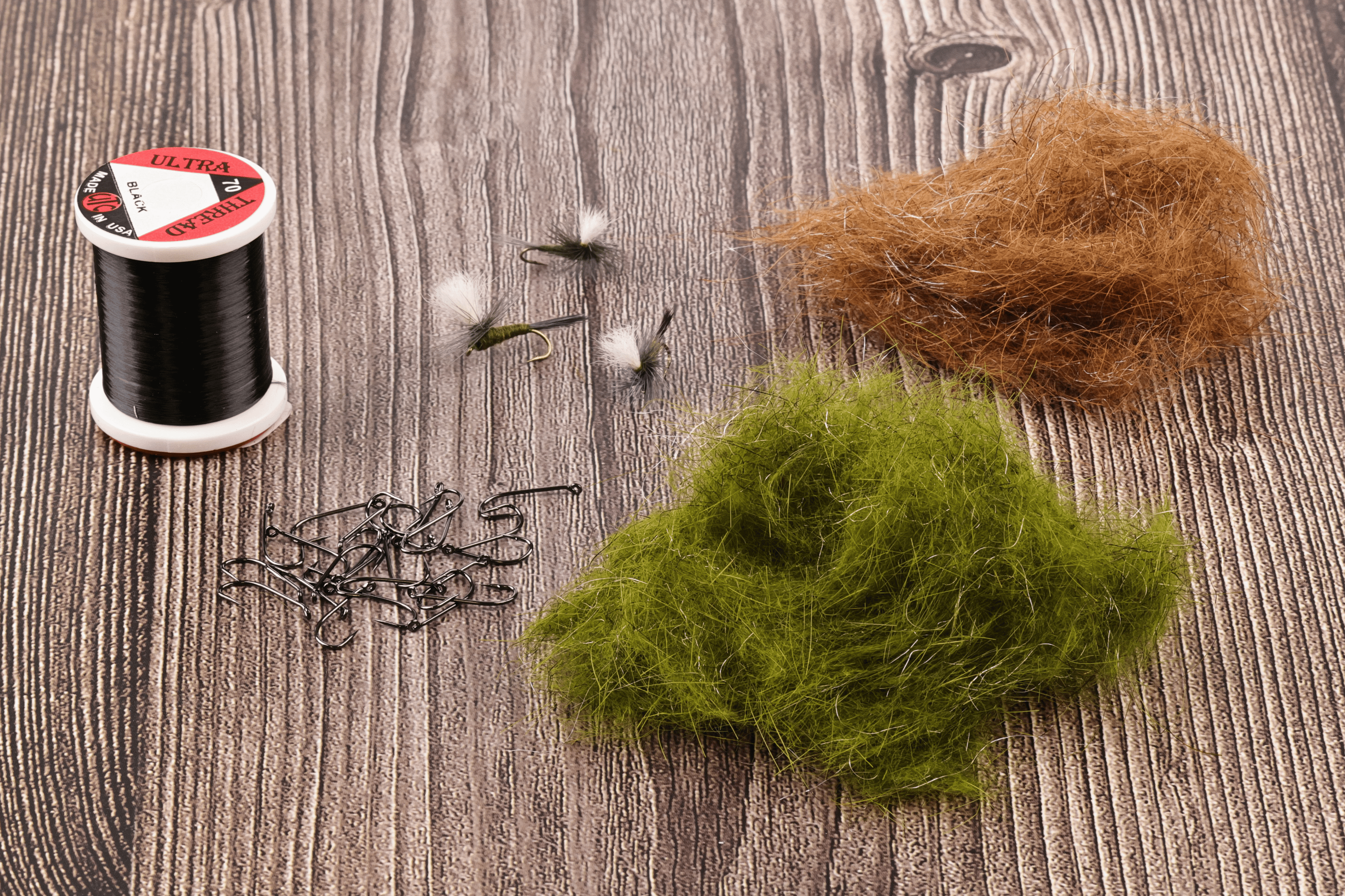Ice dubbing clumps in olive and brown with metallic fibers beside hooks and a black thread spool for fly tying