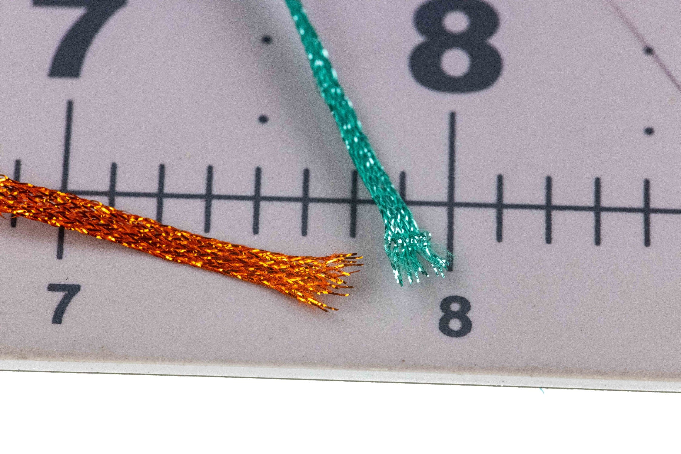 Teal and orange metallic flat fly braid strands with frayed ends on ruler, synthetic tinsel for fly tying
