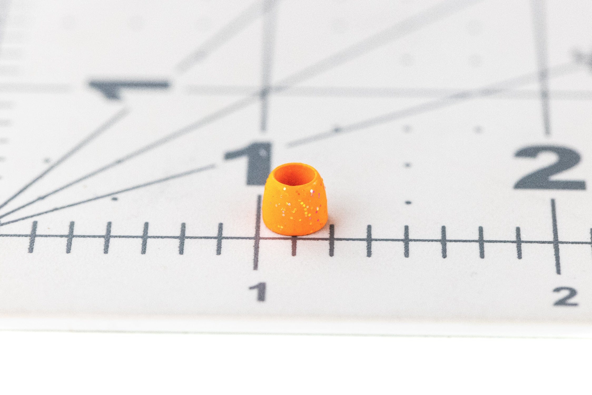 Fluorescent orange plastic tube cone head with gold fleck glitter, tapered fly tying cone placed on a measuring grid