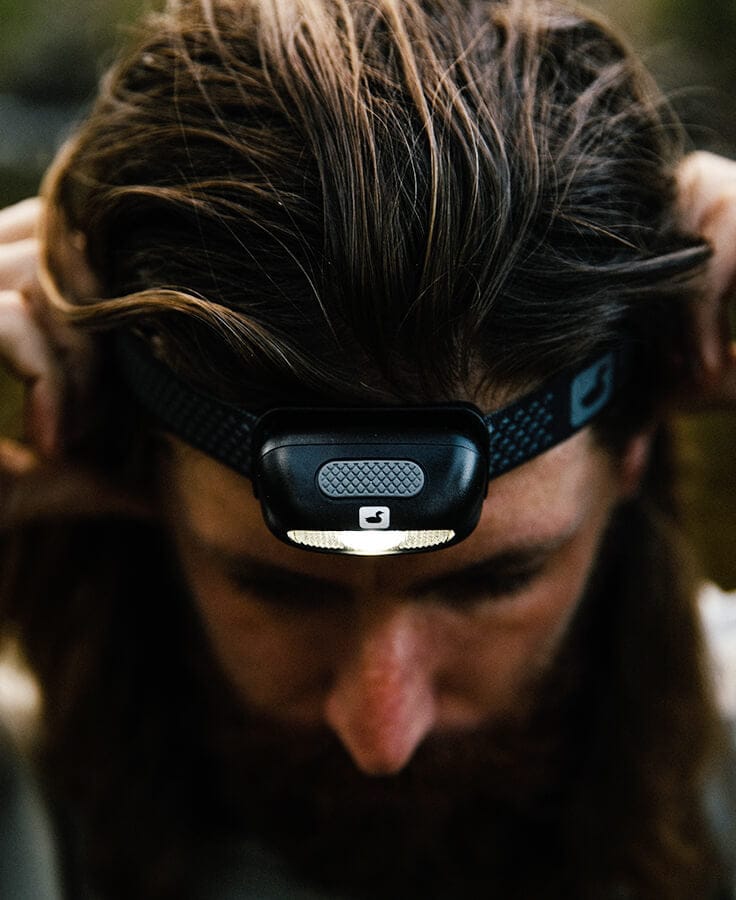Black Loon headlamp with compact rubberized LED, textured button and patterned elastic strap worn on forehead for fly tying