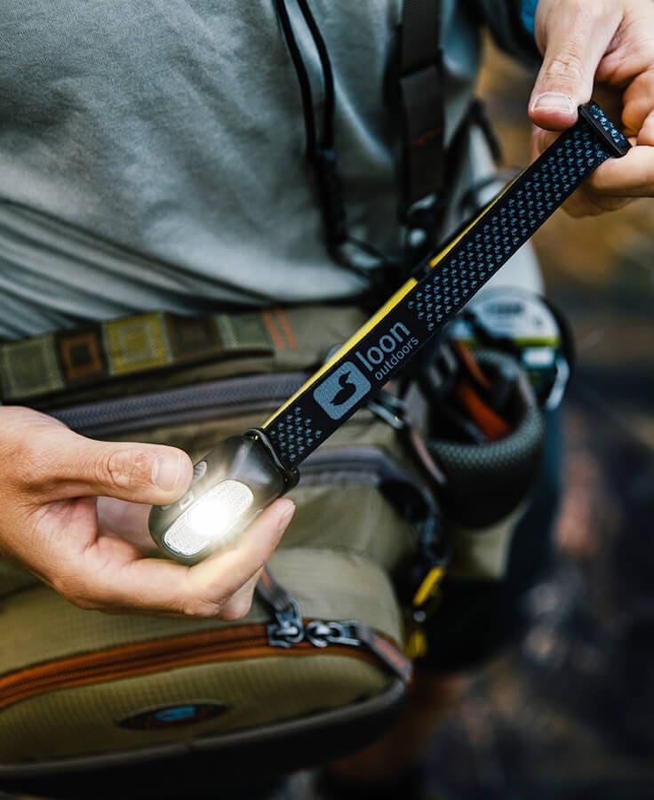 Black and yellow Loon Nocturnal LED headlamp with patterned nylon strap held over a tan fly-tying pack, light illuminated