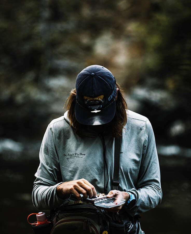 Angler in gray shirt wearing navy cap with compact black Loon Nocturnal headlamp clipped to brim while fly tying