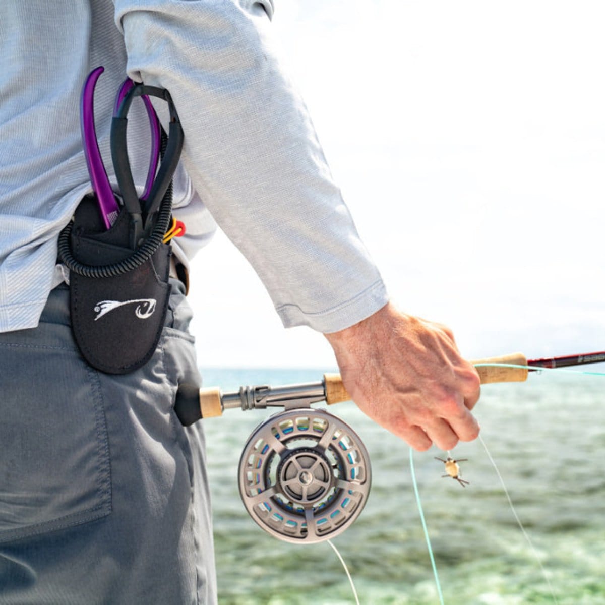 Angler's hip holster holding purple-handled fly fishing forceps with a black neoprene pouch, worn over gray pants near a