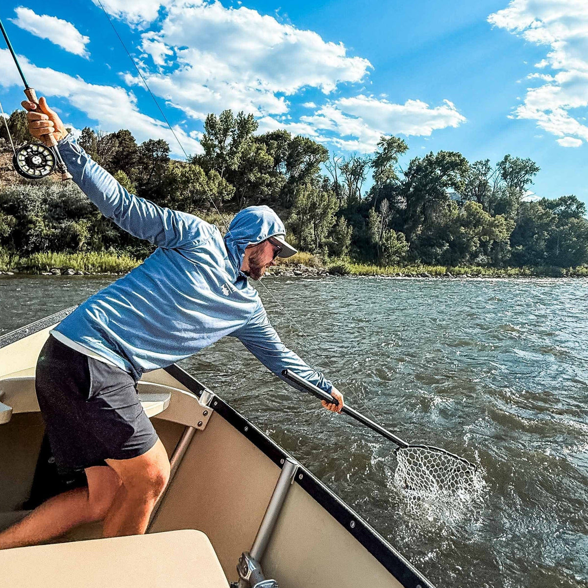 Angler in light blue hooded sun shirt uses black Rising boat net to scoop a fish from a river