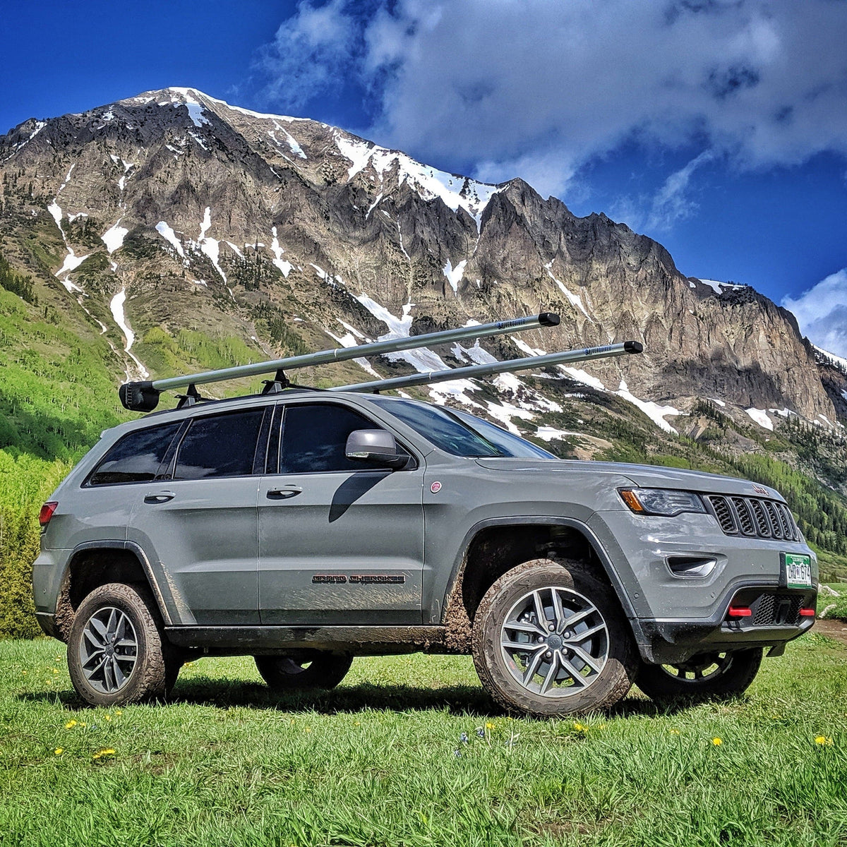 Silver Jeep Grand Cherokee with roof rack carrying Riversmith fishing rod holder in mountain landscape