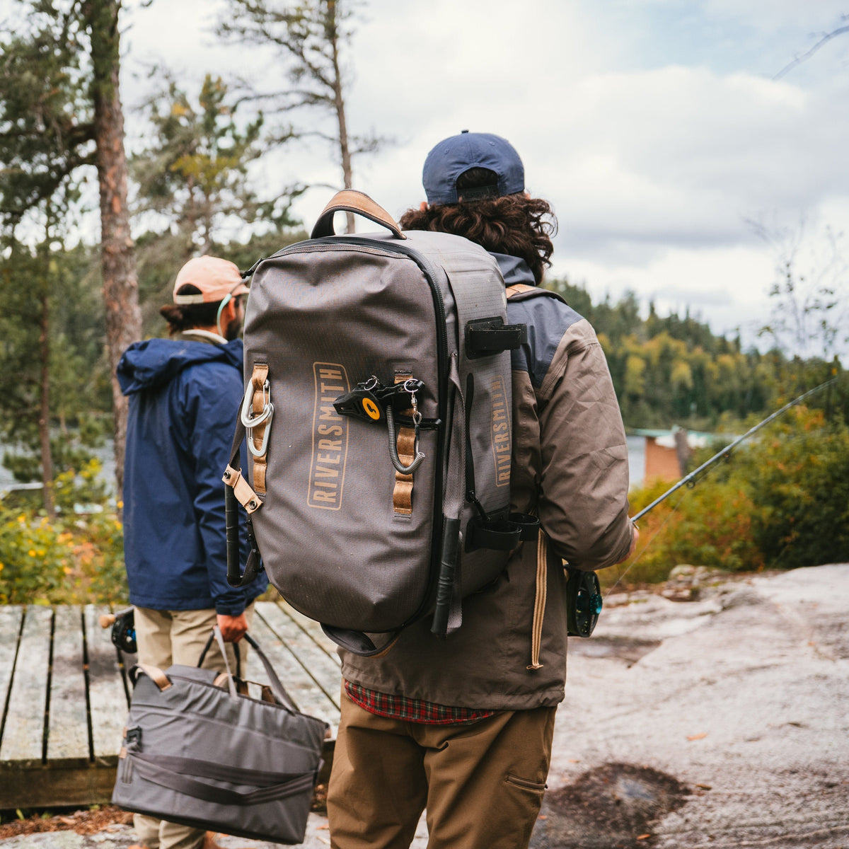 Riversmith waterproof duffel bag in charcoal gray worn by hiker with fishing gear in forest setting