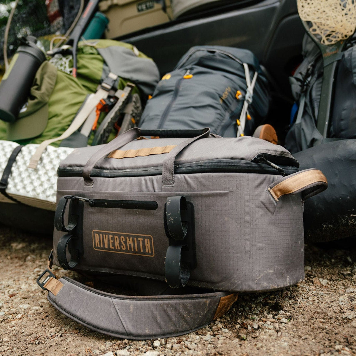 Riversmith waterproof duffel bag in gray with tan leather handles, displayed outside car with outdoor gear