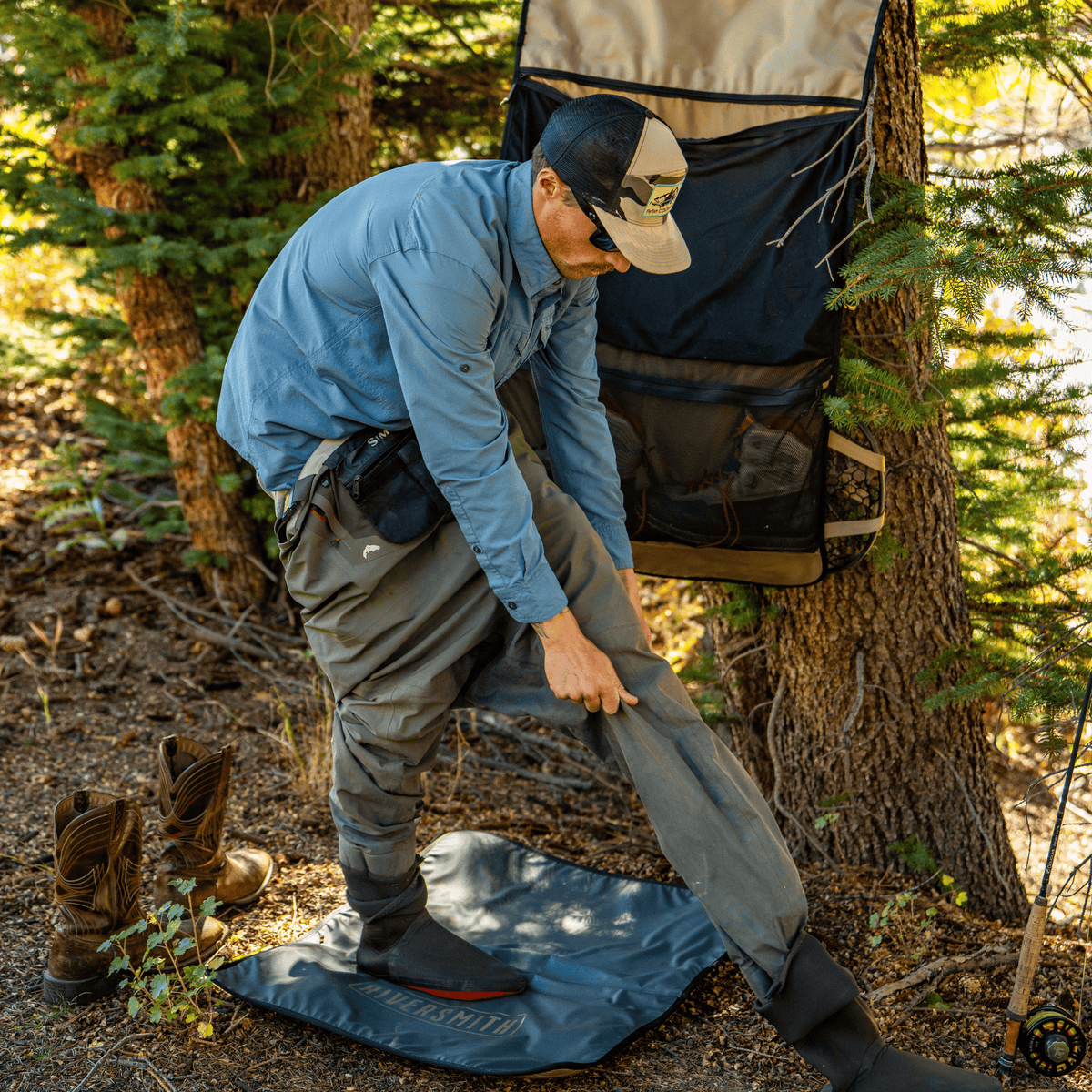 Man organizing fishing gear in blue Riversmith wader bag at campsite with trees and camping setup