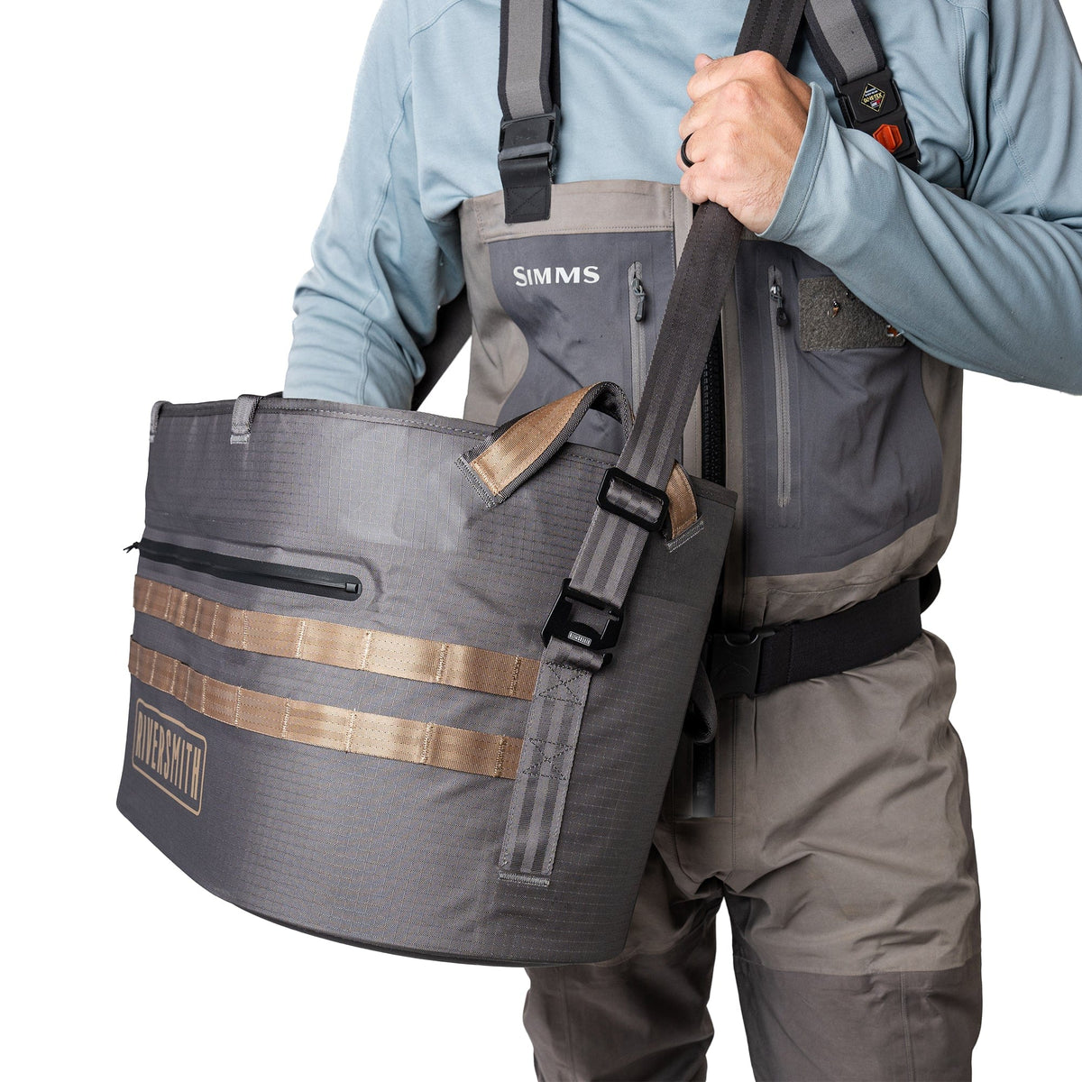 Riversmith Hauler waterproof tote bag in charcoal gray with tan straps, worn by angler over shoulder during fishing trip