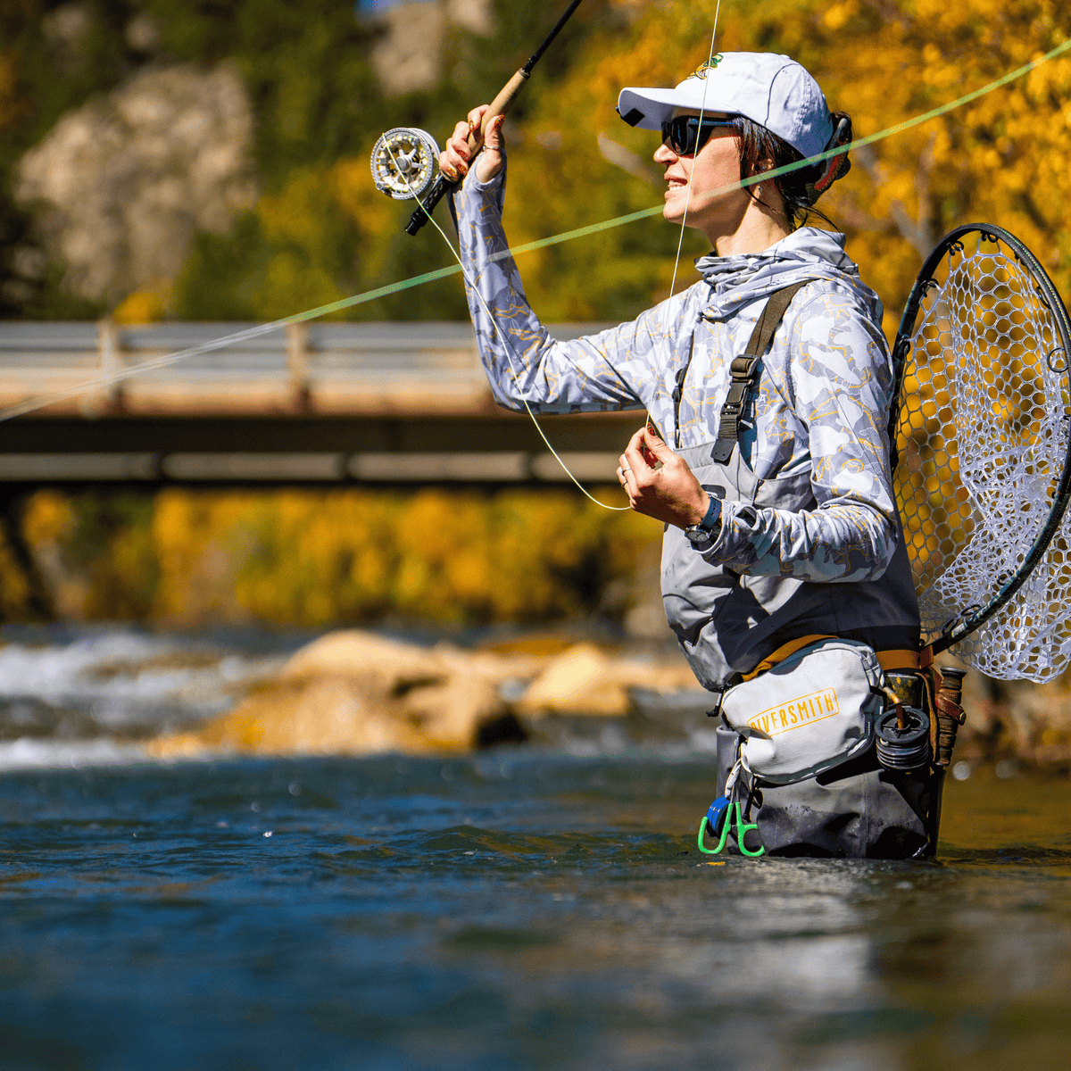 Woman fly fishing in river wearing gray Riversmith PackOut fishing pack with net and rod