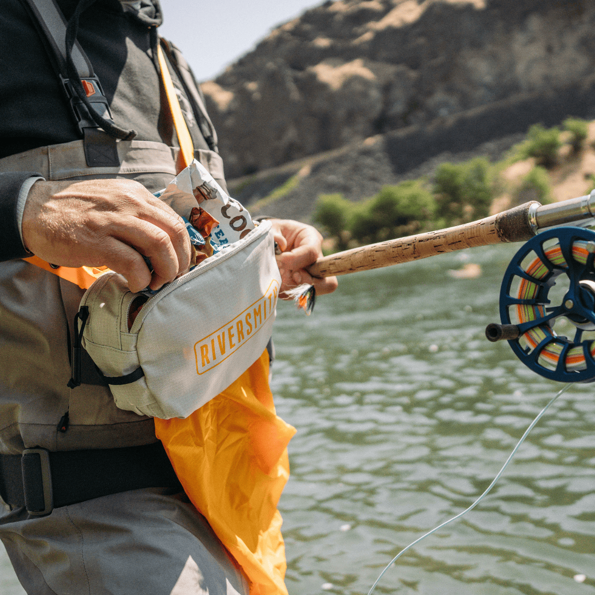 Riversmith PackOut fly fishing pack worn by angler holding fishing rod near river with mountains in background