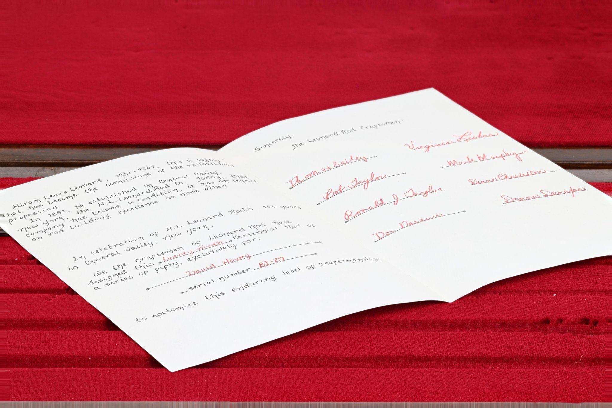 Signed white certificate with black and red cursive signatures on red ribbed fabric and a wood slat, fly rod certificate