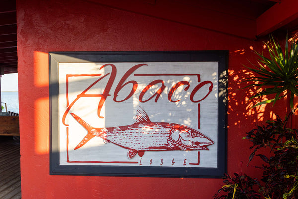 White framed sign with red fish and Abaco script on red stucco wall with tropical plant shadows Abaco Lodge
