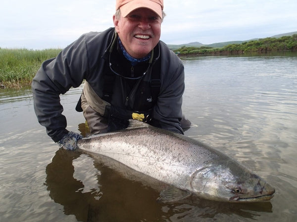 Smiling angler in gray waders and peach cap holding large silver salmon in shallow river, salmon fishing trip