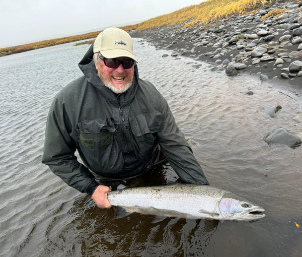 Man in dark green rain jacket and beige cap kneeling in shallow river holding large silver steelhead on rocky black sand