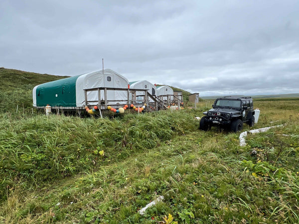 Green-and-white canvas tents on wooden platforms with colorful buoys in coastal tundra under gray sky, steelhead camp