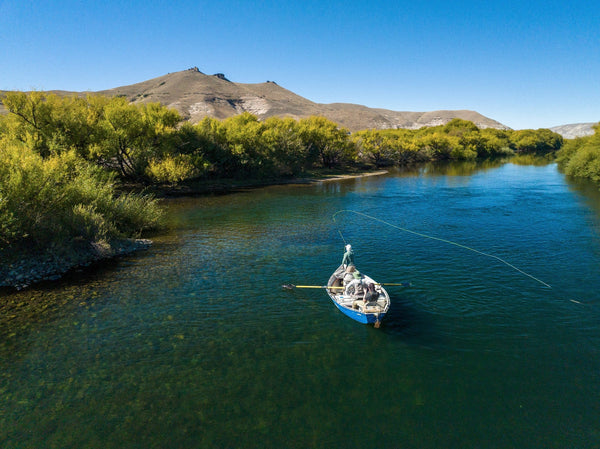 Blue drift boat with anglers fly-fishing on clear river beside willow-lined banks and brown hills under blue sky