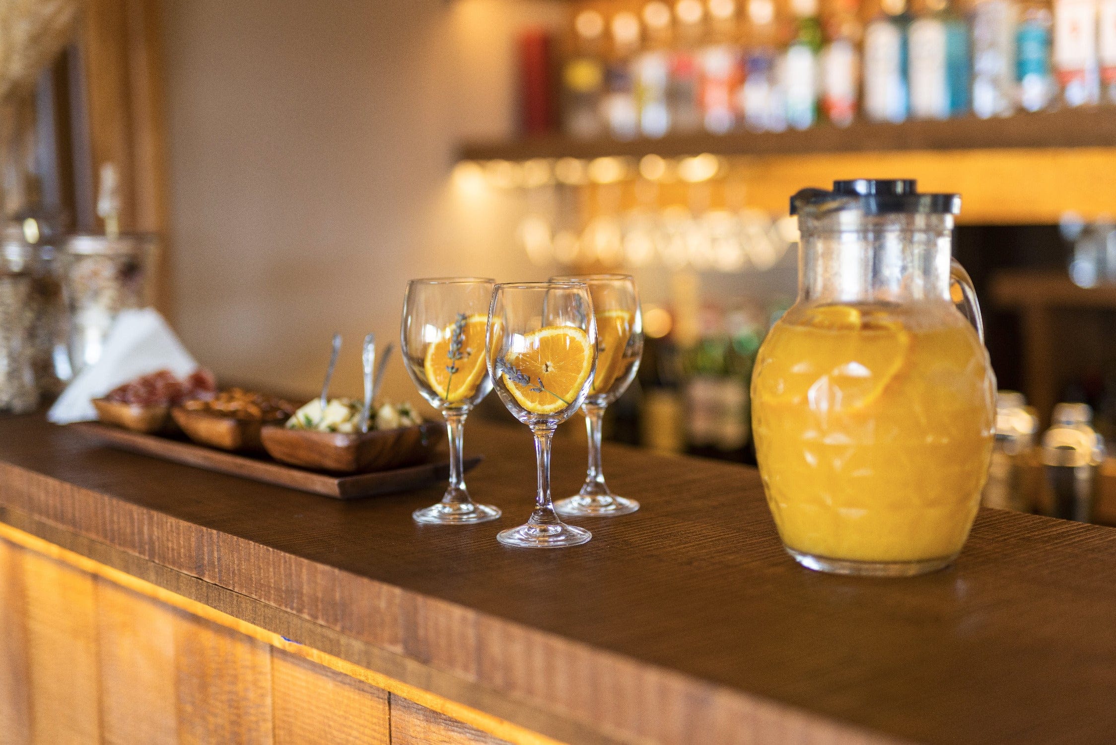 Rustic wooden bar with glass pitcher of bright orange juice and three wine glasses garnished with orange slices, safari trip