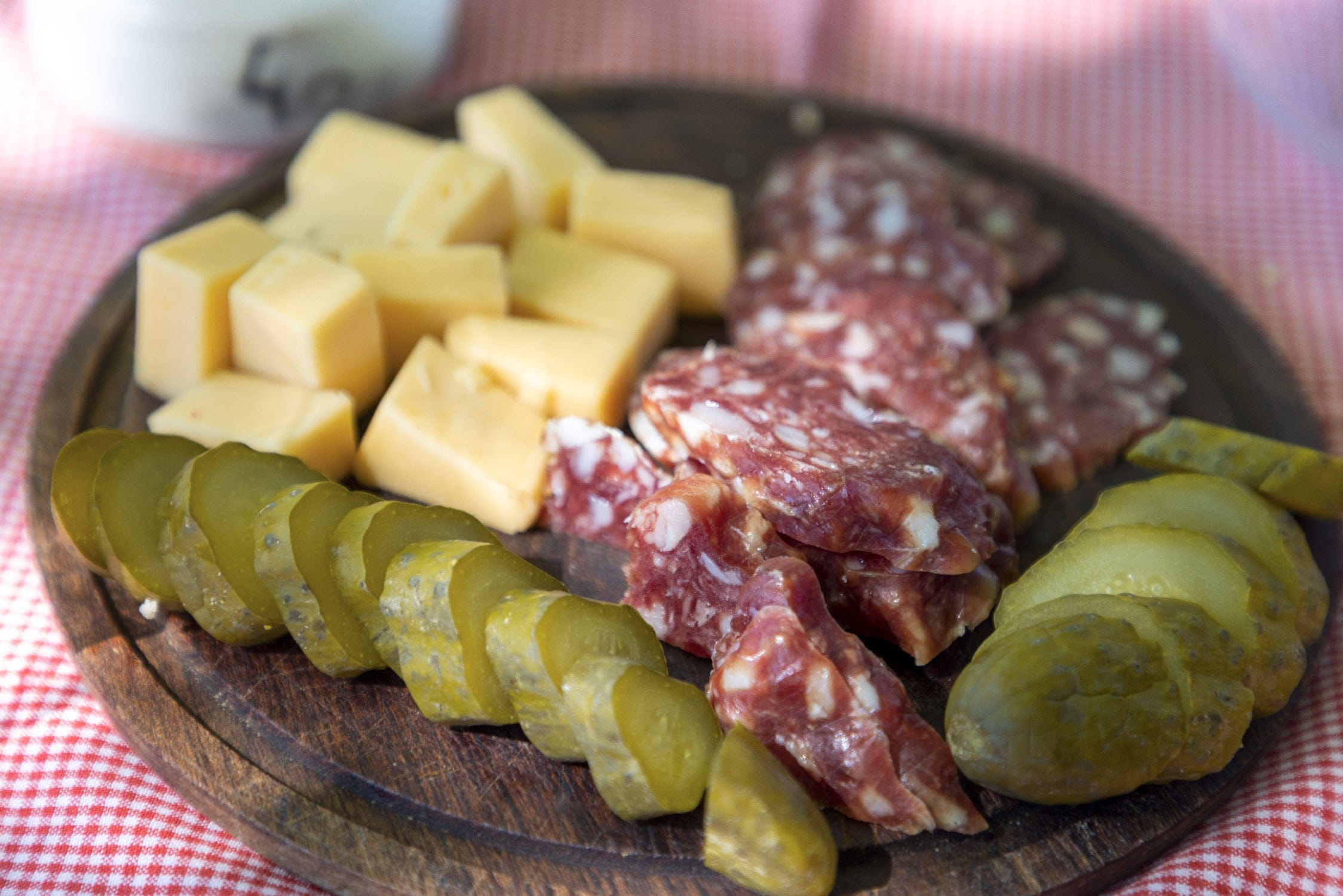 Wooden board charcuterie with yellow cheese cubes, marbled salami slices and green pickles on red gingham cloth