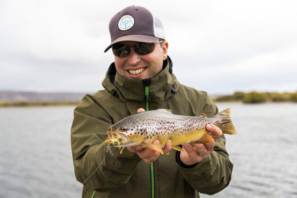 Smiling angler in olive green Gore-Tex jacket and brown Trident cap holding a speckled brown trout on a Chimehuin safari