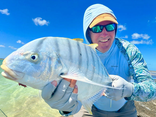 Angler in blue hoodie and teal sunglasses holding a large silver fish over turquoise shallows on a Christmas Island fishing