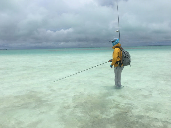 Angler in yellow jacket and gray backpack fly-fishing in turquoise shallows under cloudy sky, Christmas Island trip
