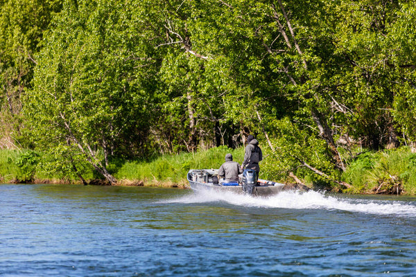 Aluminum motorboat with outboard engine and anglers speeding across blue river, wake splashing near dense green birch trees