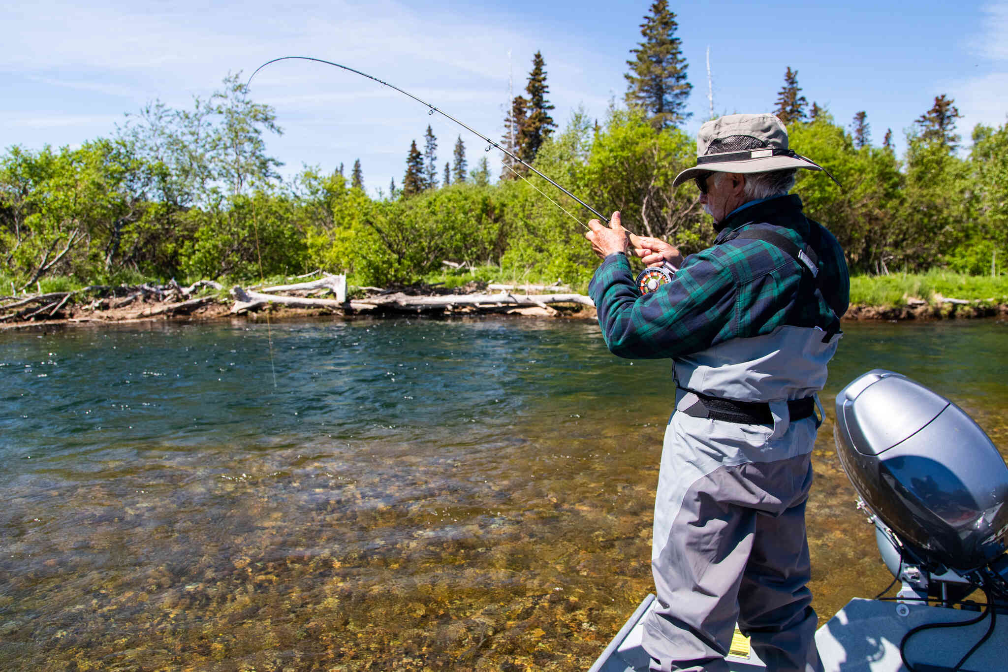 Angler in beige hat and green plaid jacket fly fishing from a boat on clear river with evergreen bank, Copper River Lodge