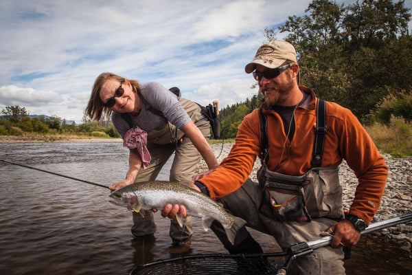 Two anglers in tan waders and sunglasses holding a silver rainbow trout at a rocky riverbank on a Copper River trip