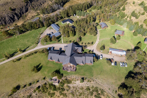 Coyhaique River Lodge aerial: dark-shingle lodge with wooden deck, gravel drive, small cabins on green lawns amid trees
