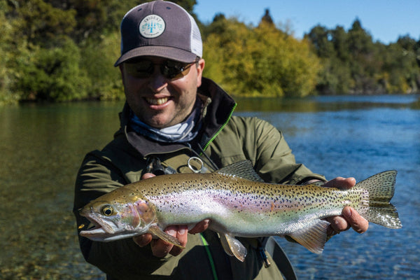 Angler in Trident cap and olive jacket holding large speckled rainbow trout on lodge trip by clear river and green trees