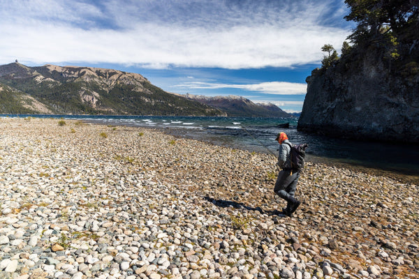 Pebble beach beside turquoise mountain lake, angler with backpack near rocky cliff — Estancia Arroyo Verde Lodge trip