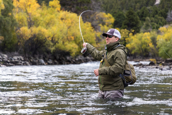 Angler in olive-green waterproof jacket and chest waders fly fishing in a river with yellow autumn trees — lodge trip