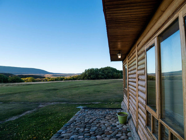 Wooden log lodge with glass windows and pebble stone porch overlooking green fields and distant hills under a blue sky
