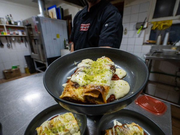 Black ceramic plate with baked rolled enchilada topped with melted white cheese and green herb crumb, held by chef in lodge