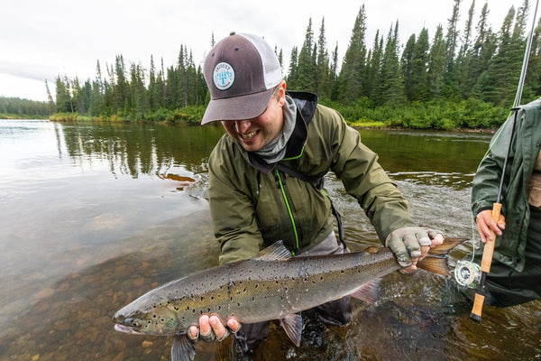 Angler in olive-green jacket and brown Trident cap holding large salmon in shallow river with evergreen trees, fly fishing