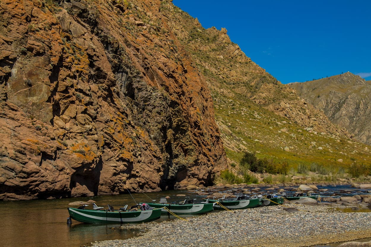 Painted green drift boats lined on pebble riverbank below towering rust-colored canyons under clear blue sky
