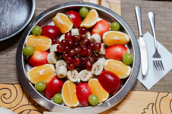 Stainless steel fruit platter with orange wedges, apple slices, banana rounds and red and green grapes; fork and knife