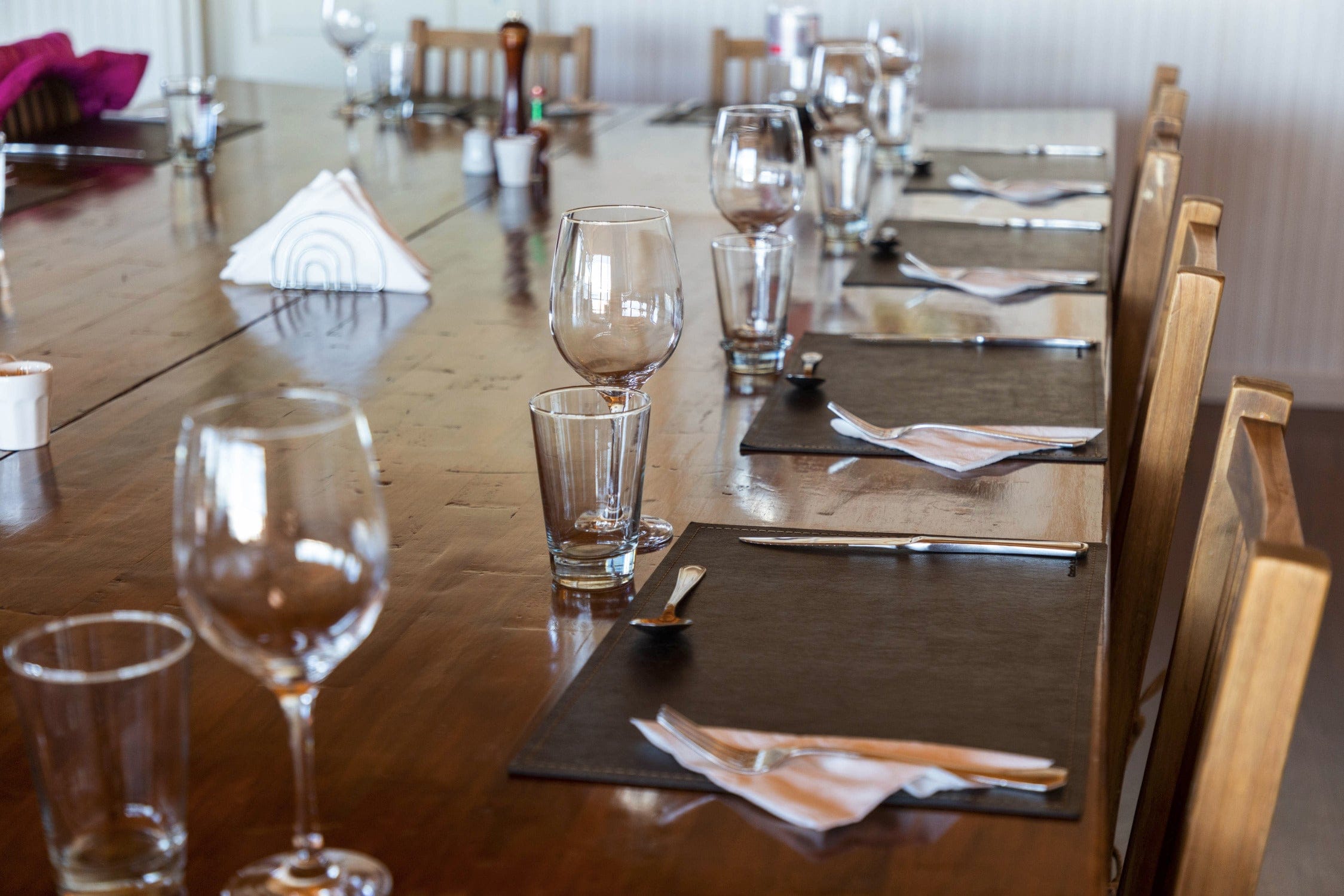 Warm brown wooden dining table with clear wine and water glasses, black placemats and folded napkins in lodge dining setup