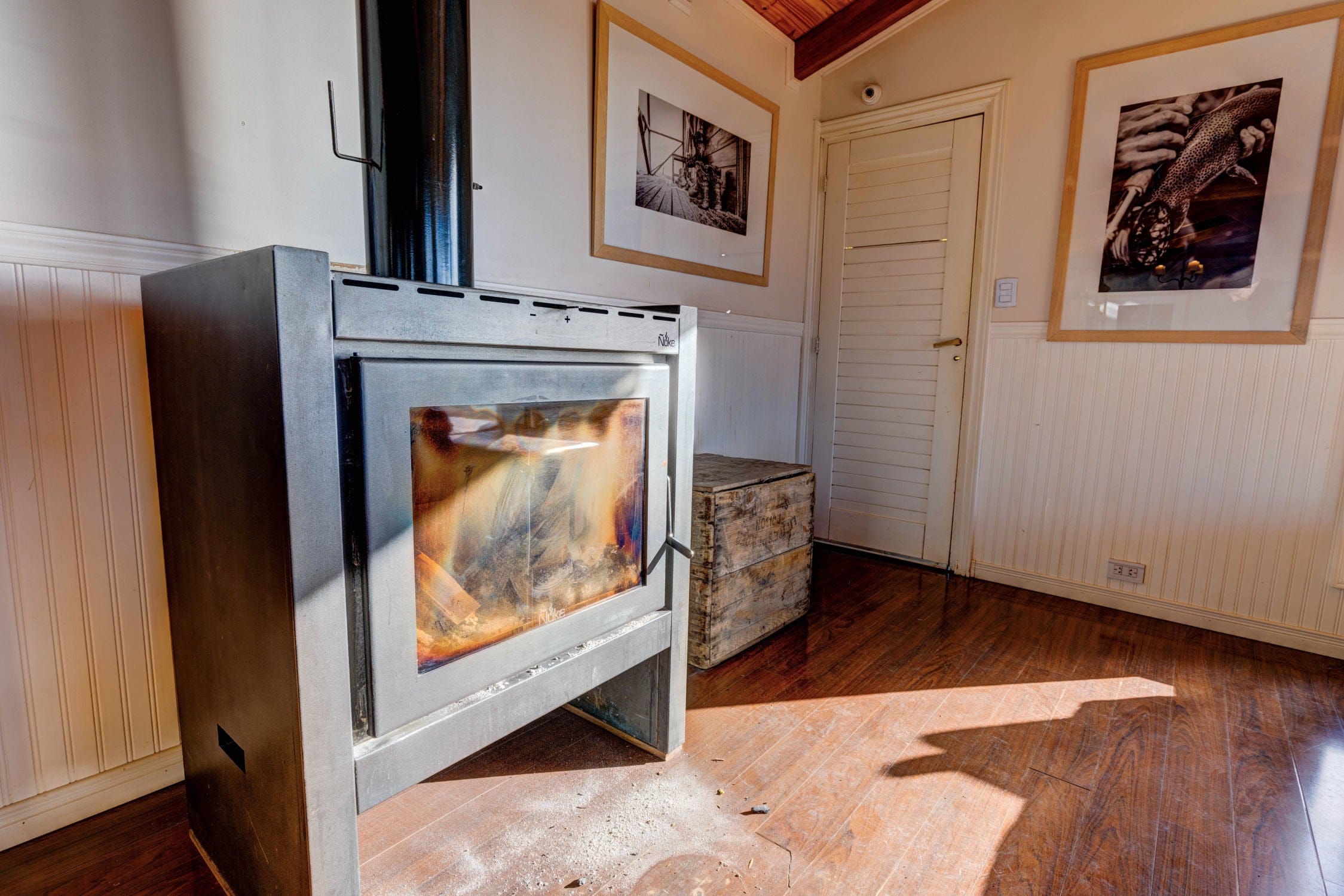 Gray metal wood-burning stove with visible flame and black flue against white beadboard walls and hardwood floor in lodge
