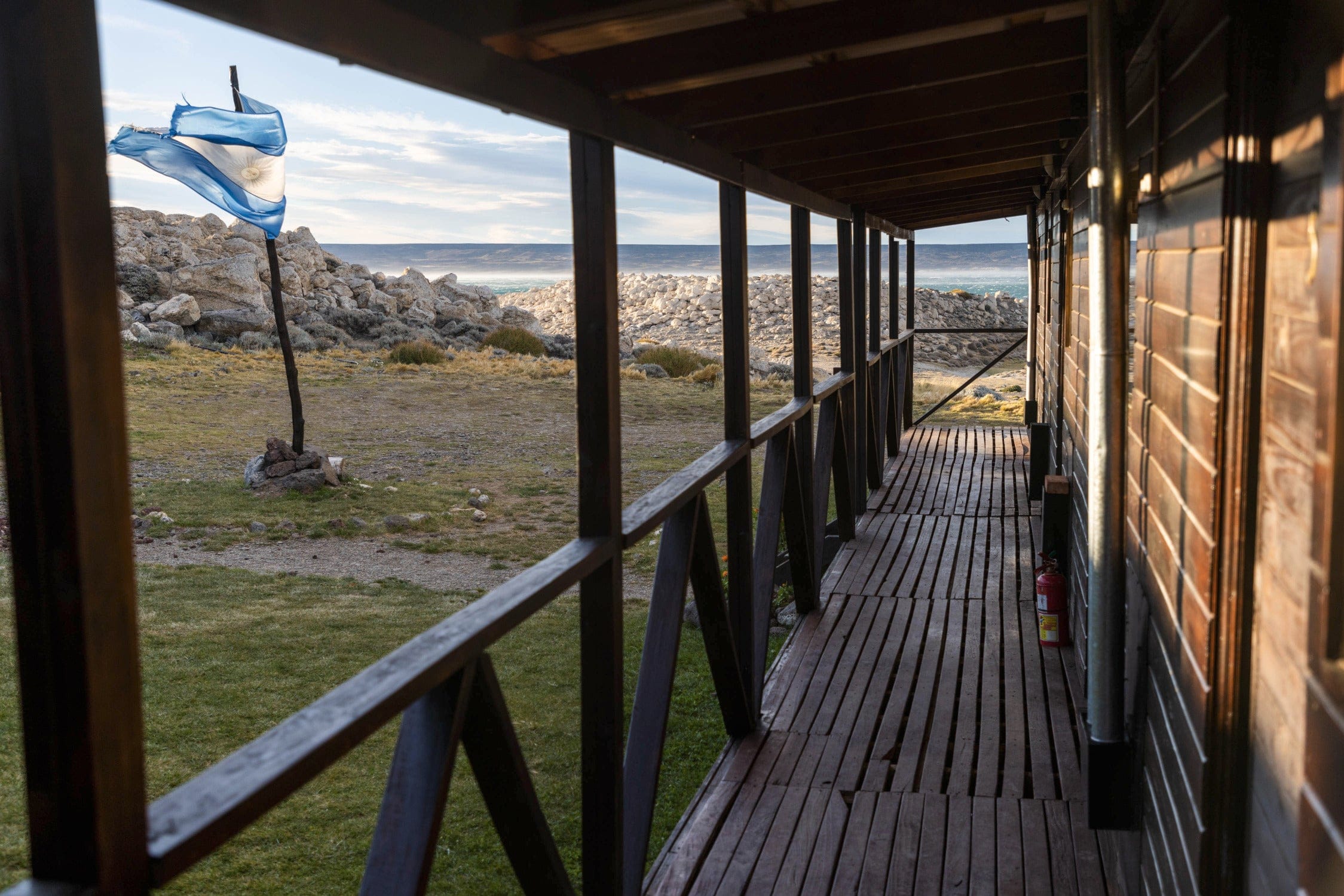 Rustic dark-wood lake lodge porch overlooking rocky shore, waving blue flag and distant ocean horizon