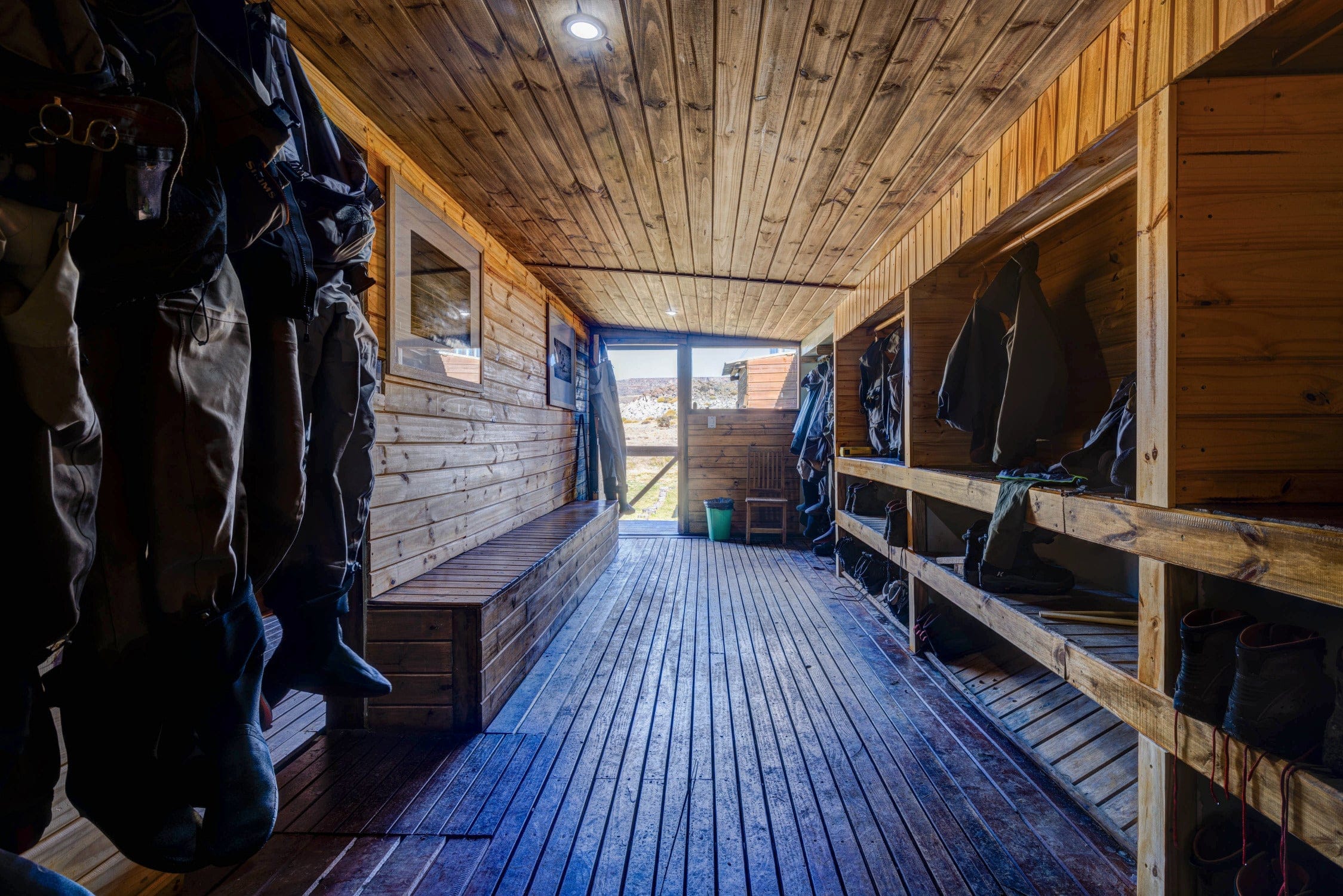 Rustic pine bootroom at Jurassic Lake Lodge for fishing trips with wood-plank floor, benches, hanging waders and boot shelves