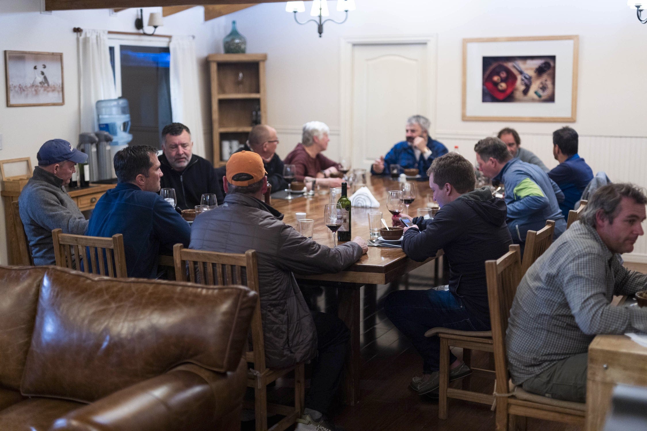 Rustic dining room with long wooden table, wooden chairs, leather sofa and guests sipping wine, ideal lodge trip.