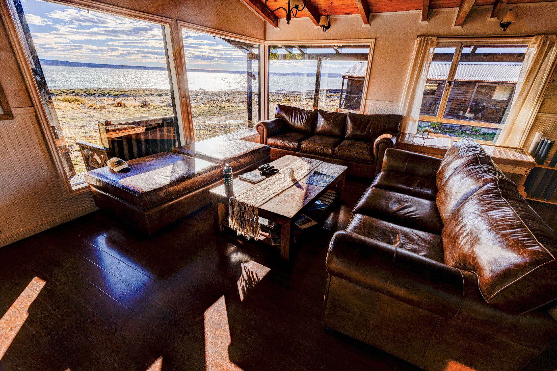 Cozy lakeside lodge living room with brown leather sofas, wooden table and large windows overlooking blue lake