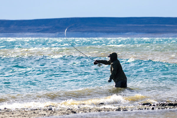 Angler in dark green hooded jacket wading turquoise lake, casting a fishing rod with arcing line against distant blue cliffs