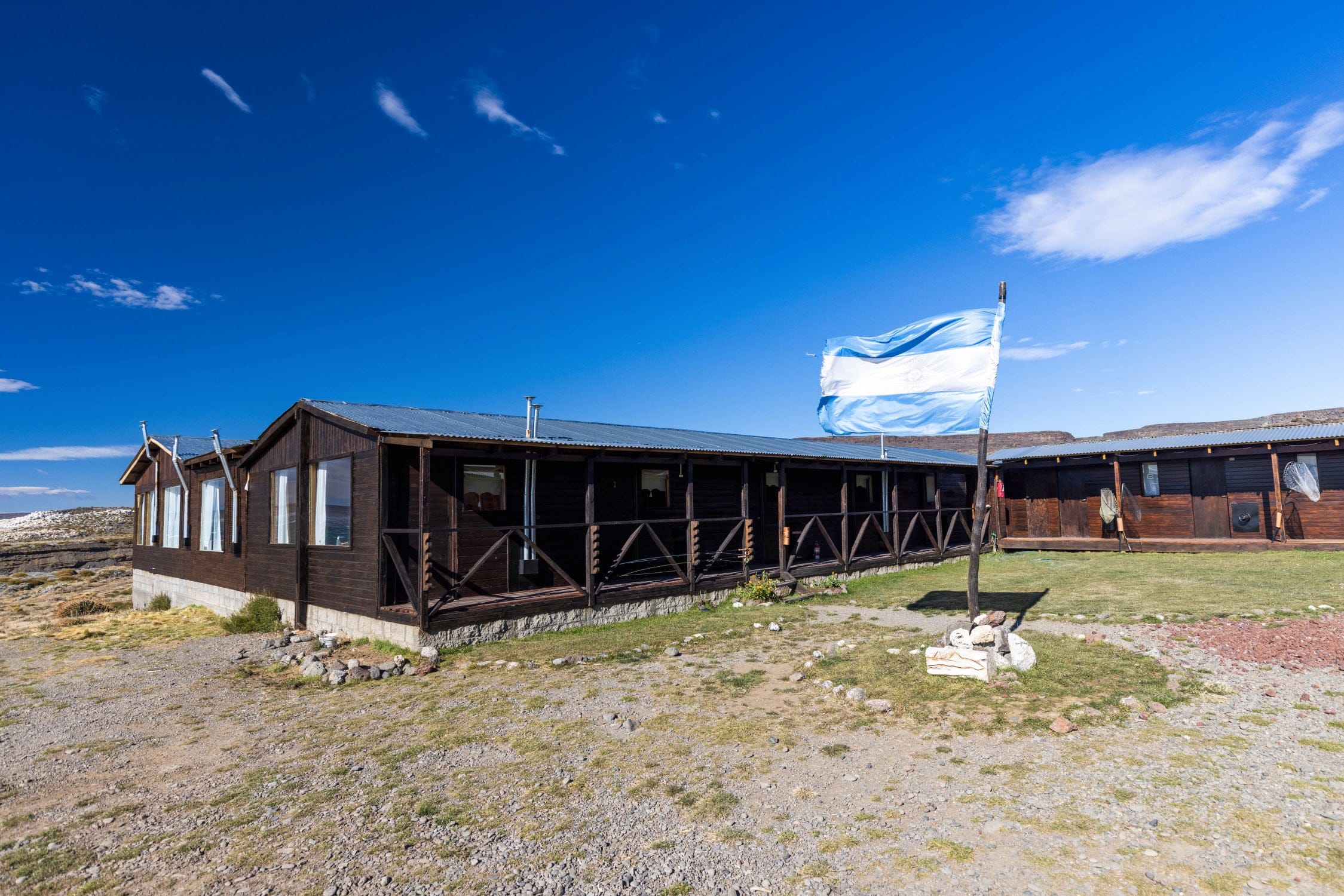 Dark wooden Jurassic Lake Lodge with corrugated metal roof and blue-white flag under vivid blue sky
