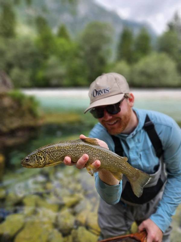 Angler in beige cap and blue technical shirt holding golden-brown trout over clear rocky river — Krka River Lodge fishing