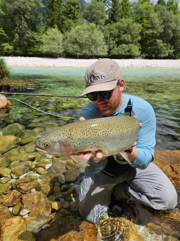 Angler in beige cap and blue shirt kneeling in clear turquoise Krka River holding large spotted trout, fly fishing trip
